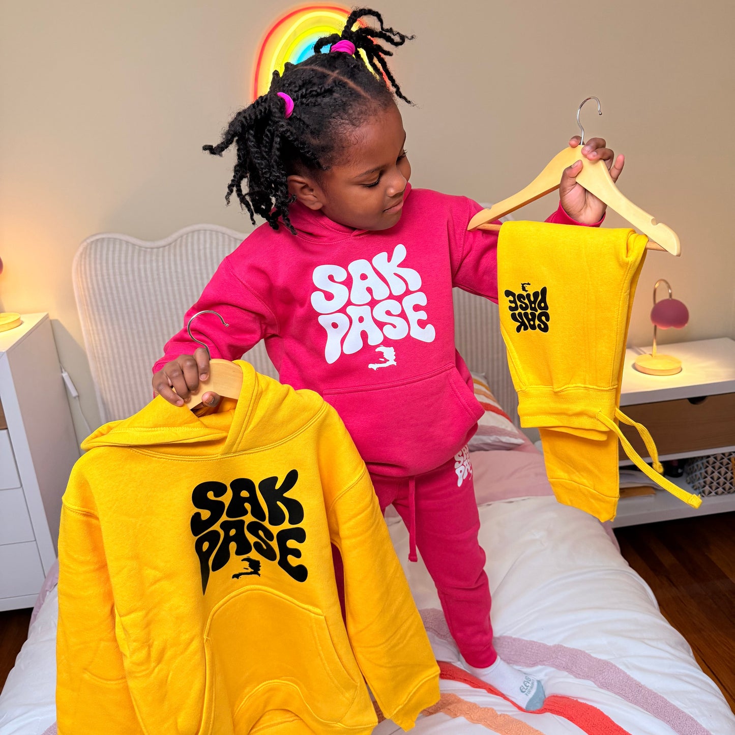 Child holding yellow and pink sweatshirts with visible branding indoors.
