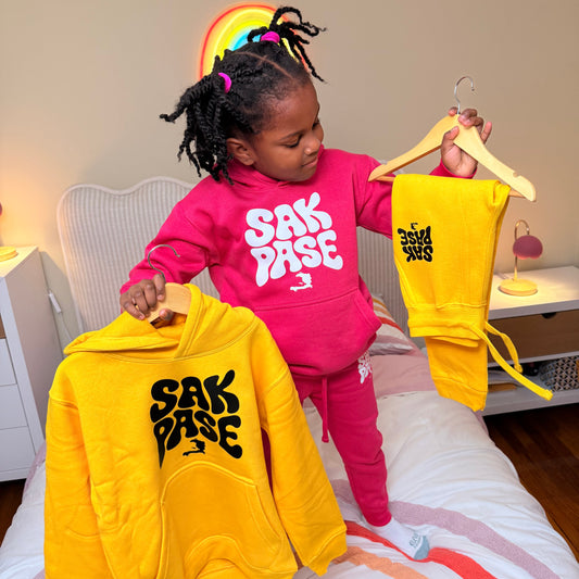 Child holding yellow and pink sweatshirts with visible branding indoors.